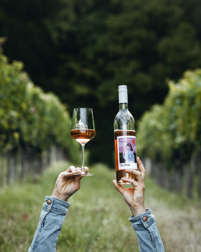 Jemand hält eine Flasche Rosi und ein Glas gefüllt mit dem Roséwein in die Luft während er im Weingarten liegt
