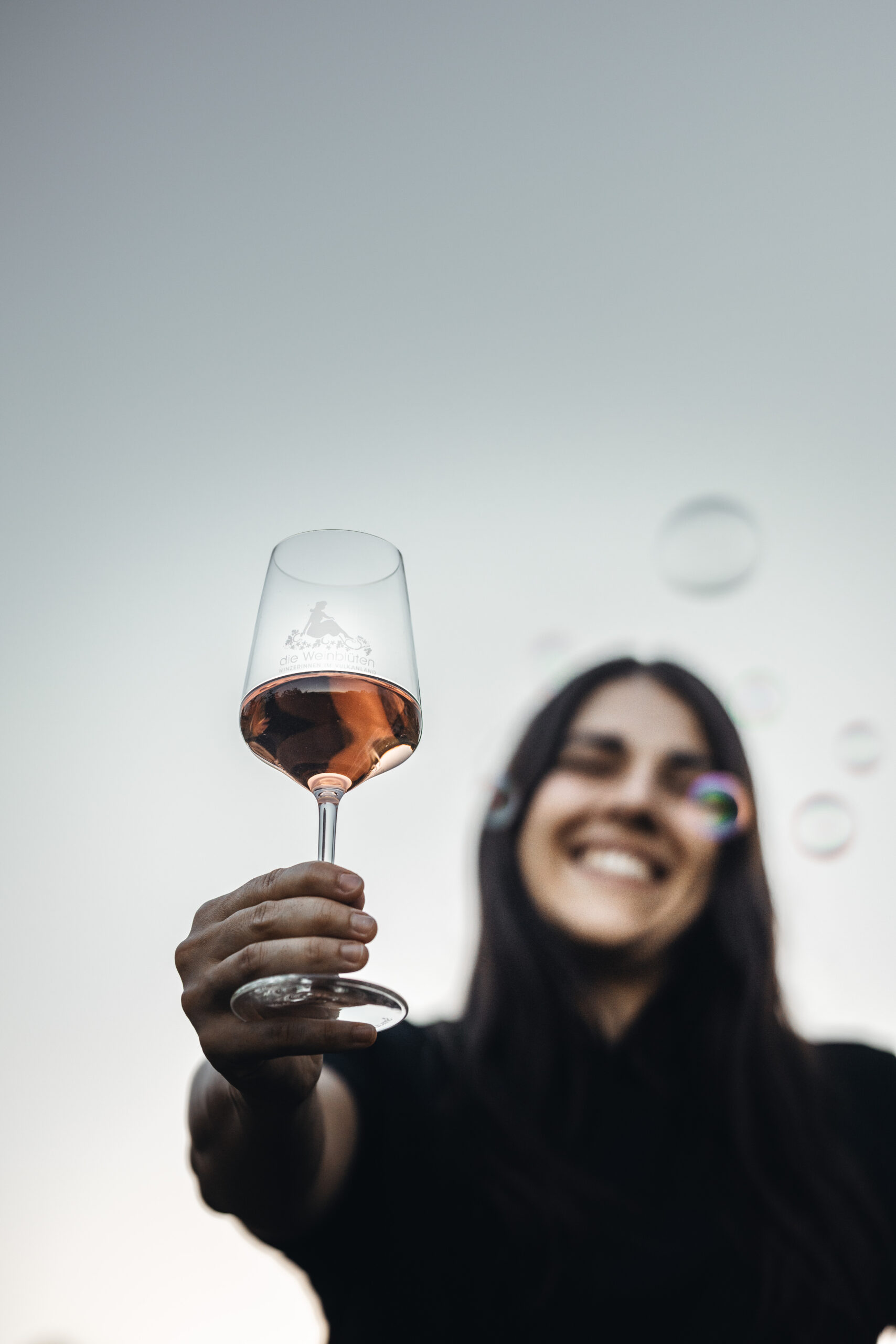 Weinblüten-Winzerin Anne Hütter-Grießbacher hält ein Glas Rosi gegen die Kamera, Luftblasen steigen auf und sie freut sich über das Weinpaket leicht und fruchtig aus dem Thermen- und Vulkanland Steiermark